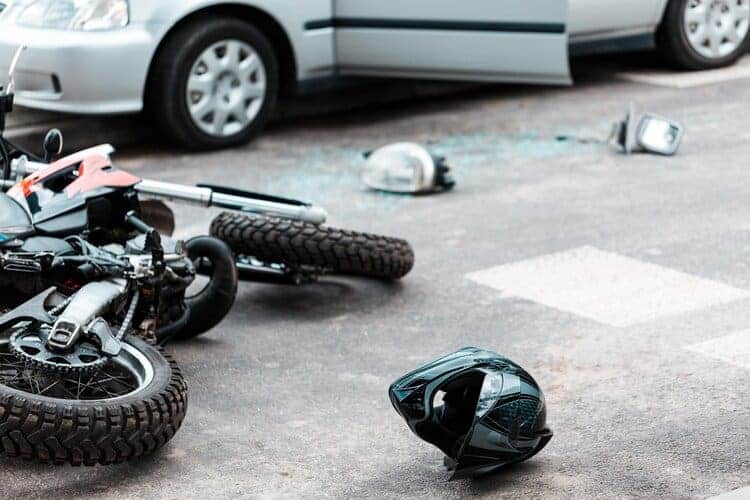 Motocicleta Tirada En El Suelo Y Casco Negro En El Asfalto Junto A Restos De Vidrios Rotos Por Un Choque Con Un Auto Plateado.