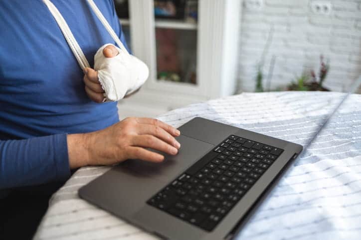Trabajador Con El Brazo Enyesado Y Cabestrillo Usando Una Laptop Para Caso De Demanda Por Incapacidad Laboral.