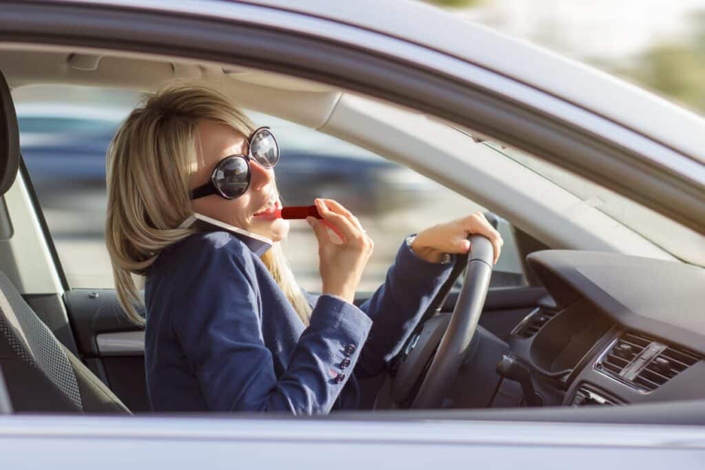 A woman multitasking while driving. She is on her phone and applying lipstick.