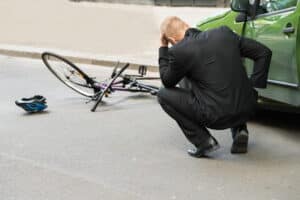 a man is kneeling next to his vehicle with his head in his hands after he hit a cyclist. The bike lays on the ground in front of the car.