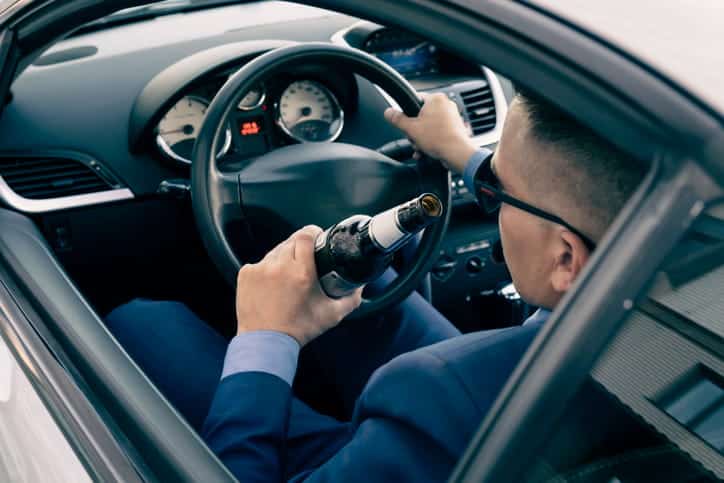 Focus is on a man behind the steering wheel of a car. He's drinking alcohol while driving.