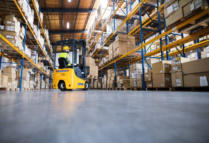 What Is The Most Common Type Of Lift Truck Accident? 1 A Photo Taken From The Ground, Looking Up At A Yellow Forklift While It Stacks Boxes In A Warehouse Aisle.