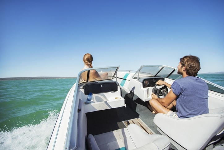 Man steering speed boat on water with woman riding up front.
