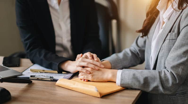 Focus is on a female lawyer with her female client. It is zoomed in on their hands as the lawyer provides comfort by placing their hand over top the clients. Underneath their hands is a manilla enveloped with documents inside.