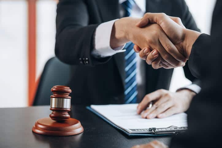 Focus is on a lawyer shaking their clients hand. In front of him is paperwork and a gavel.