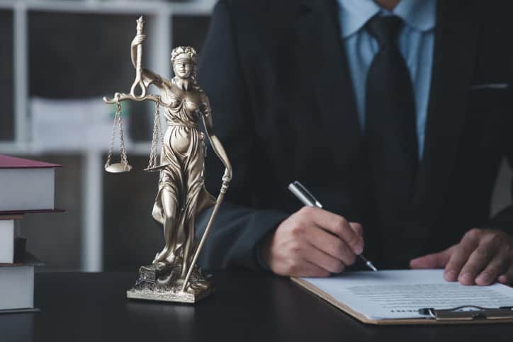 Focus is on a Lady Justice statue that is next to an unidentified lawyer working on paperwork.