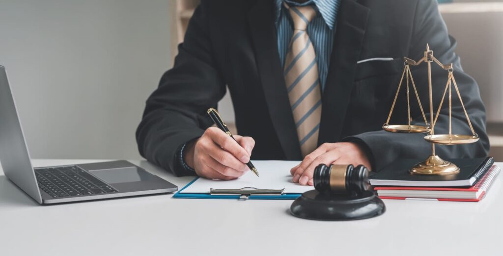 A car accident lawyer works on paperwork at his desk. Next to him is an open laptop, a gavel, and the scales of justice.