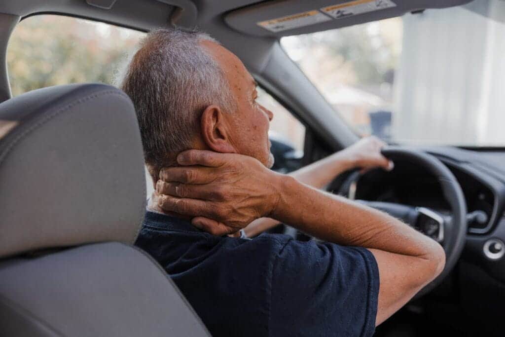 A man holding his neck in pain behind the steering wheel of a car.