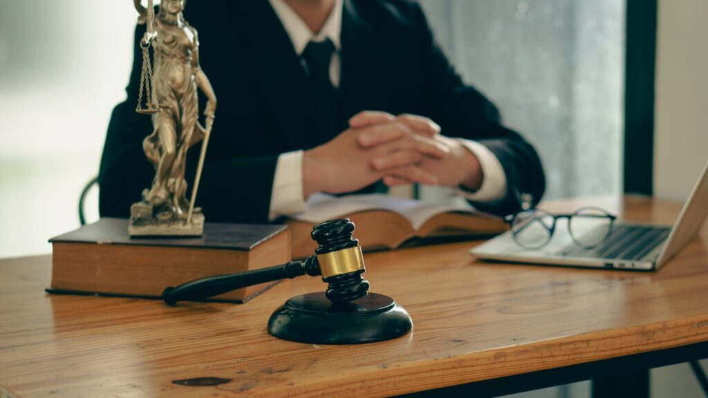 A Miami drunk driving injury lawyer sitting at their desk with their hands folded. In front of them is a book, an open laptop with their glasses resting on the keyboard, a gavel, and a Lady Justice statue. 