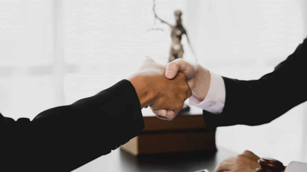 Focus is on a nursing home injury attorney shaking hands with their client. In the background is a Lady Justice statue on top of a pile of books.