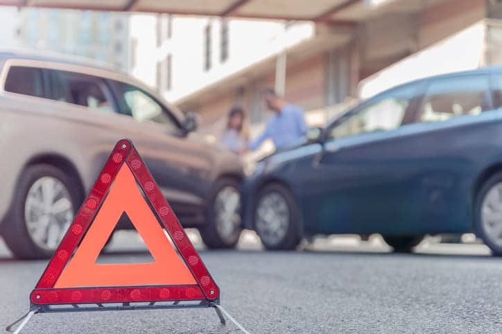 Focus is on a red caution sign warning others of a car accident. In the background out of focus is two vehicles that have collided with the drivers standing outside their cars. 