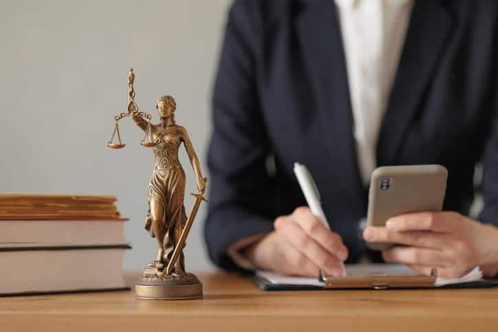 A personal injury attorney is working on paperwork as they read documents on their cellphone. Next to them is a stack of books and a Lady Justice statue. 