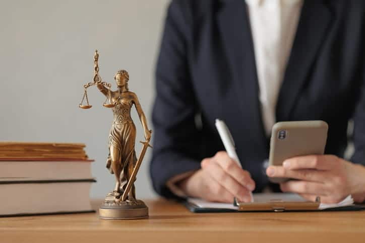 A personal injury attorney is working on paperwork as they read documents on their cellphone. Next to them is a stack of books and a Lady Justice statue.