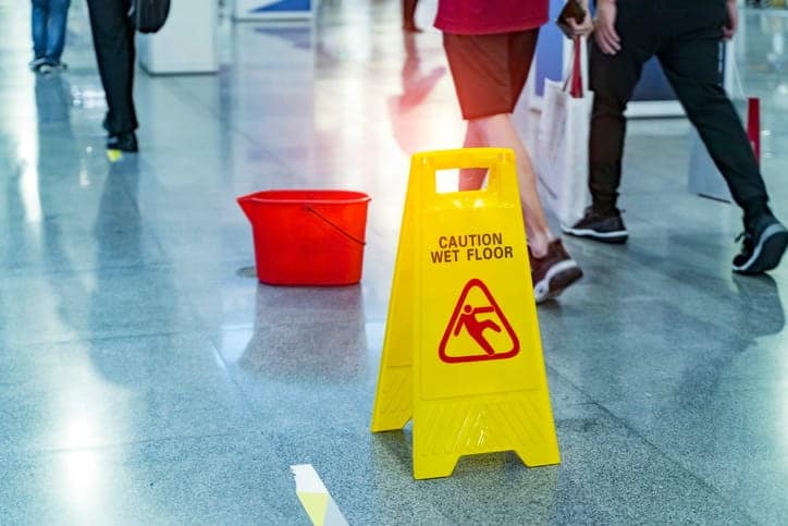 People walking by a yellow caution wet floor sign next to a red bucket.