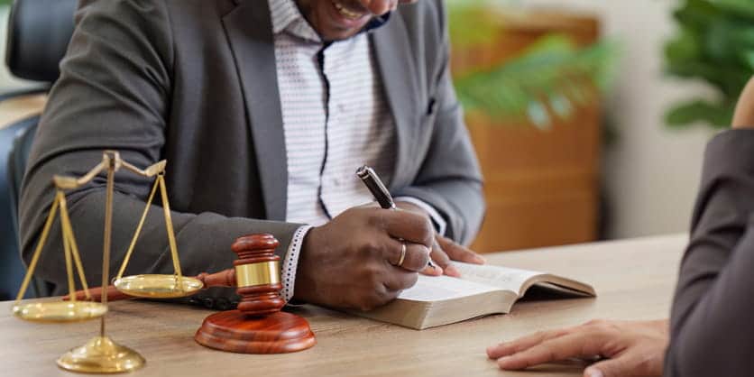 A Personal Injury Attorney Writing In A Book As He Speaks With A Client. Next To Him Is A Gavel And The Scales Of Justice. 