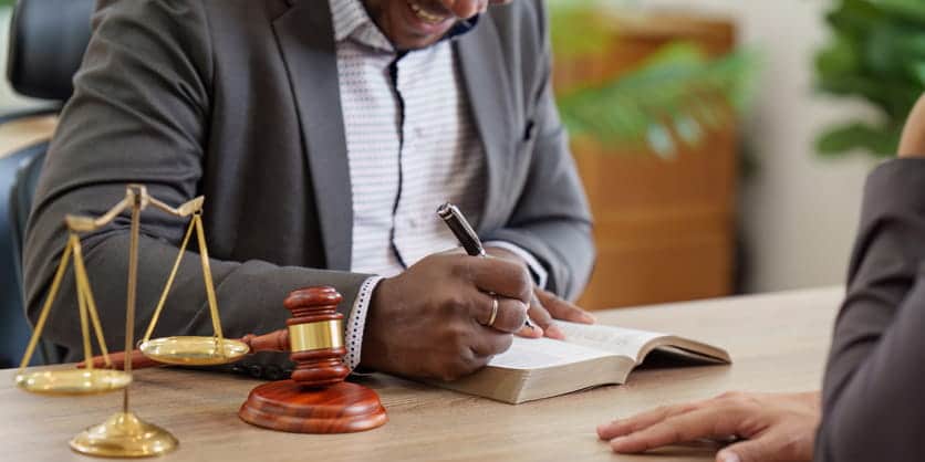 A personal injury attorney writing in a book as he speaks with a client. Next to him is a gavel and the scales of justice. 