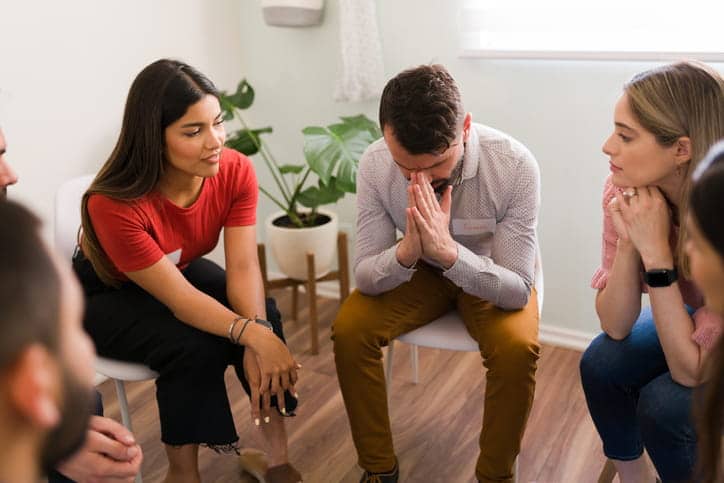 A group sits in a circle as they discuss addiction. The man in the center has his face in his hands.