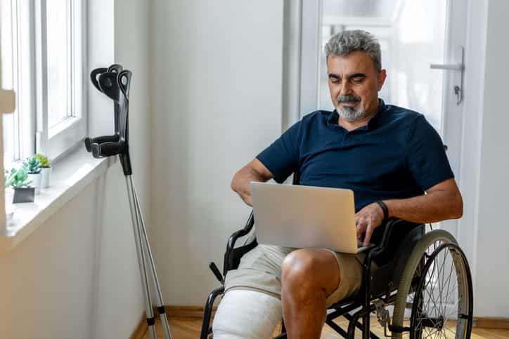 A man is in a wheelchair with an arm crutch next to him. He's on his laptop searching for a personal injury attorney after an accident.