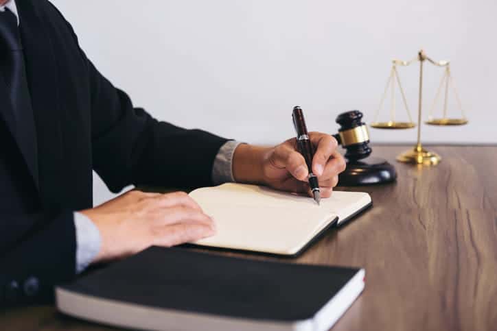 A medical malpractice attorney writing in a notebook at this desk. Next to him is the scales of justice, a gavel, and a book. 