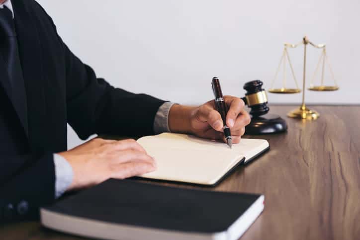 A Medical Malpractice Attorney Writing In A Notebook At This Desk. Next To Him Is The Scales Of Justice, A Gavel, And A Book. 