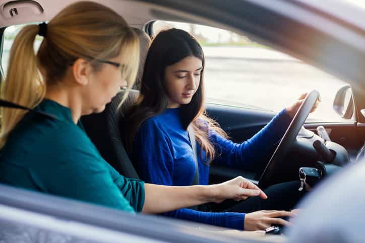 A mother showing her teenage daughter different controls behind a steering wheel of a car. 