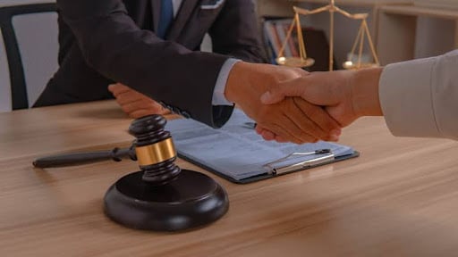 What Happens If You’re Caught Driving Without Insurance In Florida? 1 A Lawyer Shaking Hands With Their Client. On Their Desk Is A Gavel, Paperwork, And The Scales Of Justice.