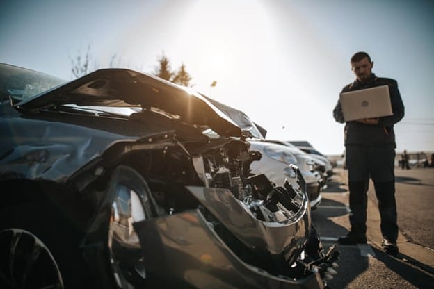 Car Accident Insurance In Florida 1 A Car Accident Insurance Scene Showing A Damaged Gray Car With An Open Hood And An Inspector Holding A Laptop, Assessing The Damage On A Sunny Day.