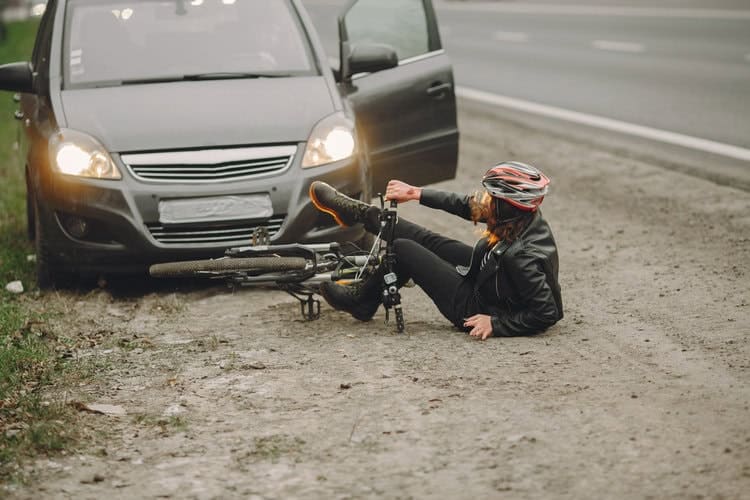 A Cyclist In A Helmet Fallen On The Ground Next To A Bicycle After Colliding With A Car On A Roadside.
