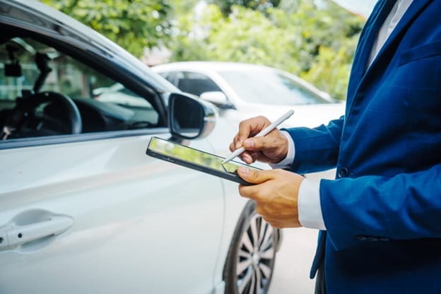 An Inspector In A Suit Using A Tablet To Assess A Car For A Property Damage Claim, Standing Next To A White Vehicle In An Outdoor Setting.