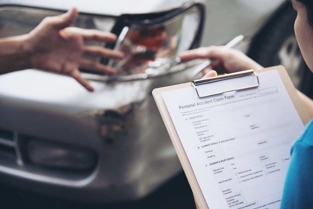 Person Holding Accident Claim Form Near Damaged Car While Another Gestures At The Damage

