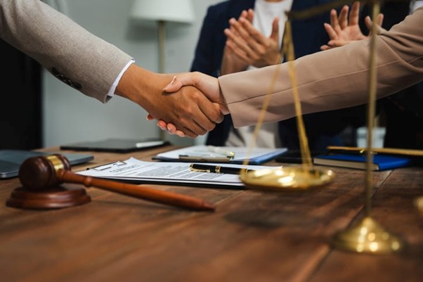 Two People Shaking Hands Over A Wooden Desk With A Gavel, Scales Of Justice, Legal Documents, And A Laptop, While Others Applaud In The Background.