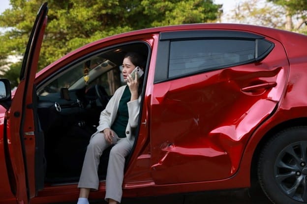 Leased Car Accidents 1 A Woman Sitting In The Open Driver’s Door Of A Damaged Red Leased Car, Speaking On A Phone With A Concerned Expression, Surrounded By Green Trees In The Background.
