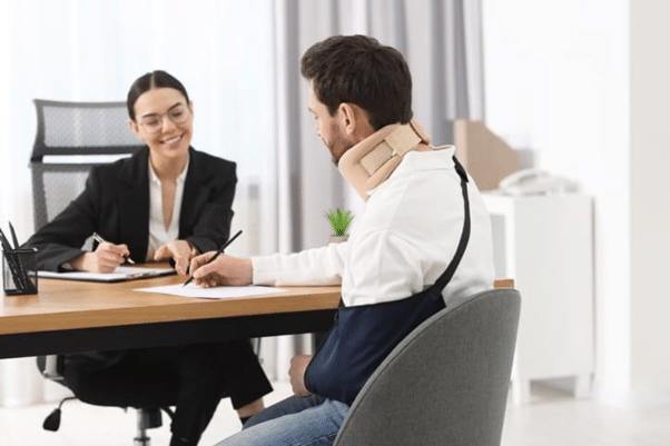 Palmetto Bay Personal Injury Lawyer 76 A Woman In A Black Suit Writing At A Desk While Consulting With A Man Wearing A Neck Brace, Both Reviewing Papers In A Modern Office With A Plant And Furniture.