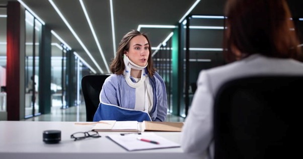 A Woman In A Black Suit Consulting With A Man Wearing A Neck Brace And Arm Sling, Seated At A Wooden Desk With Papers And A Plant In A Modern Office.