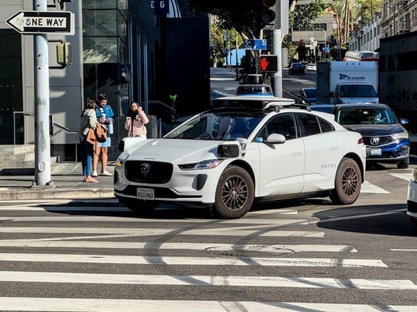 A White Waymo Self-Driving Jaguar I-Pace With Roof-Mounted Sensors And Lidar Stopped At A City Intersection, With Pedestrians Waiting At The Crosswalk And Modern Buildings In The Background.